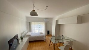A modern hotel room with a bed, a glass dining table, white chairs, a TV, and a ceiling light.