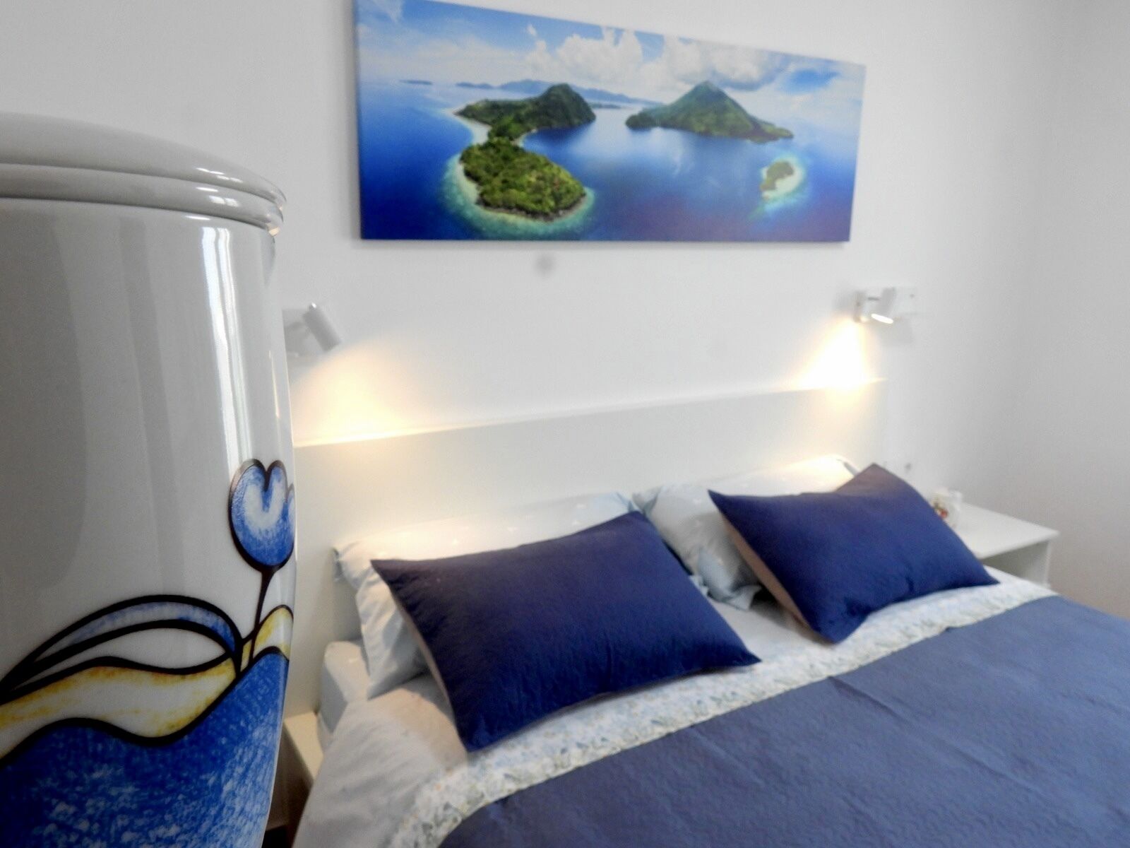 A hotel room with a bed, two pillows, a nightstand, and a painting of islands on the wall.