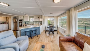 A modern kitchen with a central island, a dining area with a table and chairs, a living room with a sofa, and large windows offering a view of the landscape.