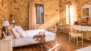 Une chambre avec un mur en pierre, un plafond en bois, un lit avec une literie blanche, un lustre, une coiffeuse avec un miroir et une petite table avec une chaise.