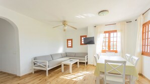 Een woonkamer met een bank, een eettafel met stoelen, een plafondventilator en een aan de muur bevestigde televisie.