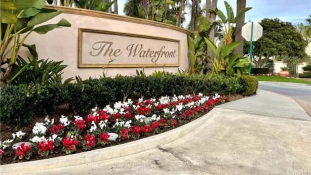 A landscaped entrance with a sign reading "The Waterfront," surrounded by tropical plants and flowers. - Coastal Condo - Gated, Pool, Spa, Walk To The Beach (Huntington Beach)