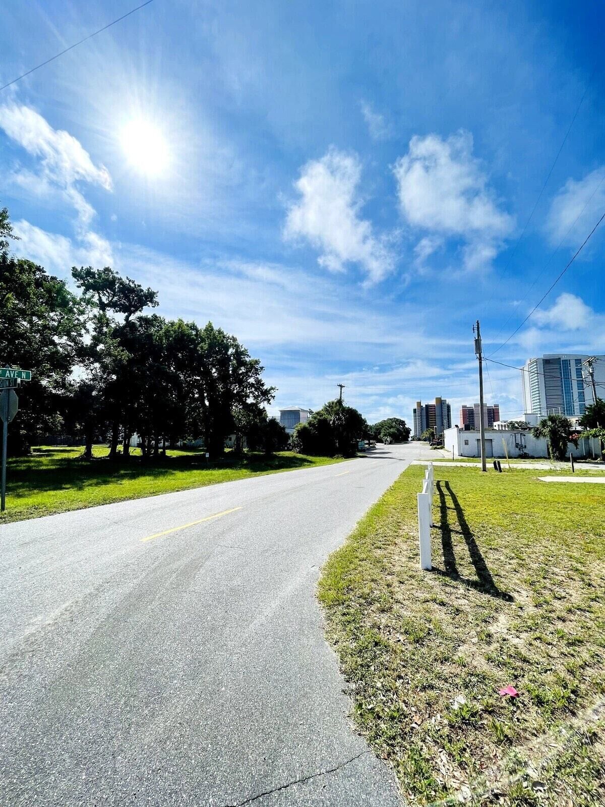 Une rue bordée d’un ciel dégagé, d’une zone herbeuse et de bâtiments visibles au loin.