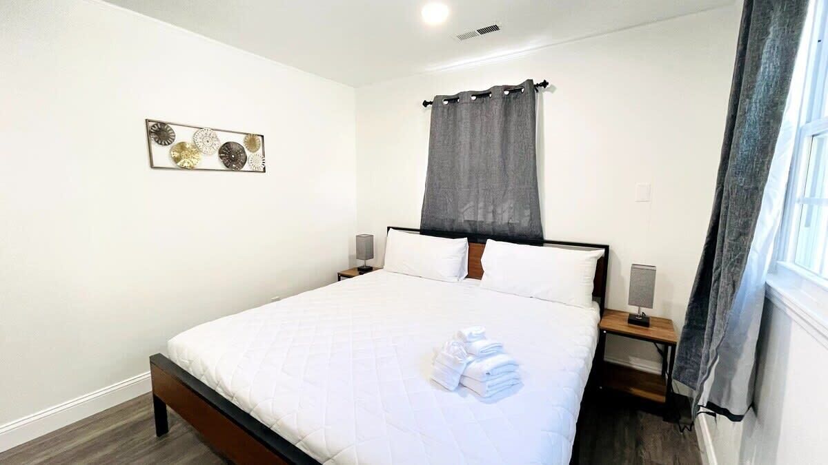 A neatly made bed with white linens and a folded towel, a bedside table with a lamp, a wall-mounted artwork, and a window with a curtain.