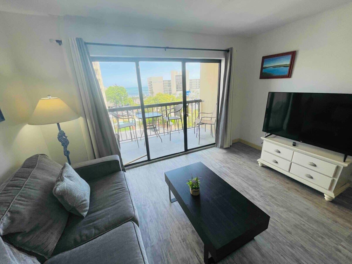 A modern living room with a sofa, a coffee table, a TV stand, and a balcony with a view of buildings and trees.
