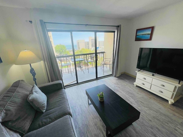 Una sala de estar moderna con un sofá, una mesa de centro, un mueble para televisión y un balcón con vistas a edificios y árboles.