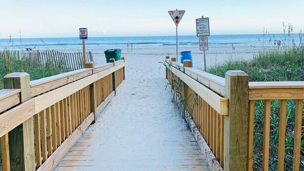 Ein hölzerner Steg führt zum Strand, mit Schildern, die eine Geschwindigkeitsbegrenzung von 24 km/h anzeigen und vor den Dünen warnen.