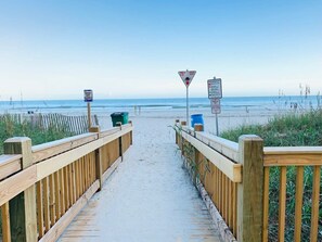 Un muelle de madera que conduce a la playa, con señales que indican un límite de velocidad de 24 kilómetros por hora y una advertencia sobre las dunas.