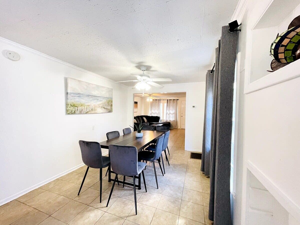 Un espace repas avec une longue table et des chaises, un ventilateur de plafond et une cuisine en arrière-plan.