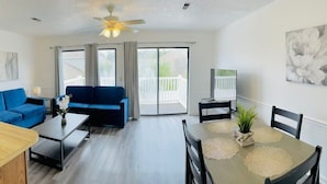 A modern living room with a blue sofa, a dining table with a placemat, and a ceiling fan.