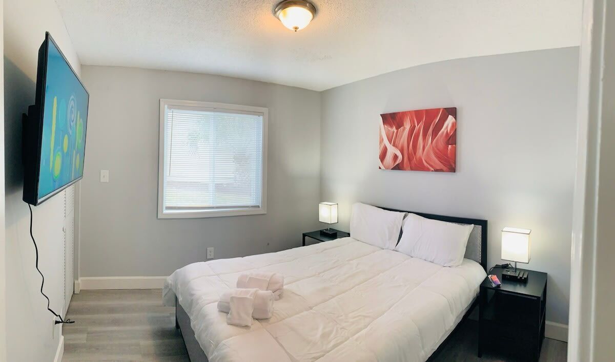 Una habitación de hotel ordenada con una cama grande, dos mesitas de noche con lámparas, un televisor de pantalla plana en la pared y una ventana con persianas.
