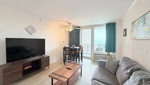 A living room with a flat-screen TV, a wooden coffee table, a sofa, a dining table with chairs, and a balcony with a view. - Carolina Winds #5 beautiful Ocean view condo, pool (Myrtle Beach)