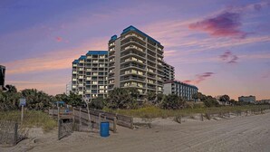 A multi-story hotel with balconies, a beach in the foreground, and a sunset sky. - Carolina Winds #5 beautiful Ocean view condo, pool (Myrtle Beach)