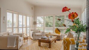 Sala de estar luminosa com grandes janelas, um sofá, uma mesa de centro e um vaso com flores.