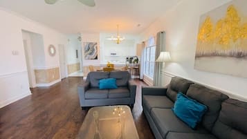 A spacious living room with a dark wood floor, two grey sofas with blue cushions, a coffee table, and a chandelier.