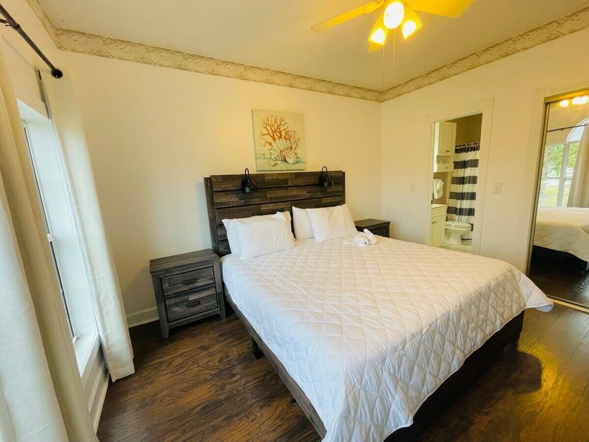 A bedroom with a large bed, a nightstand, a wooden headboard, a ceiling fan, and a painting on the wall.