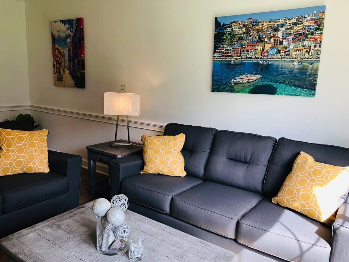 A living room with a grey sofa, two yellow patterned pillows, a small side table with a lamp, and a painting of a coastal town.