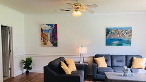 Un salon avec un canapé noir, un canapé gris et une table basse en bois. On y trouve un ventilateur de plafond et deux tableaux accrochés au mur.