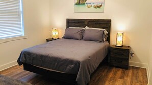 Une chambre à coucher avec un lit, des tables de chevet, un plancher en bois, une fenêtre avec des stores et un tableau représentant une scène de plage.