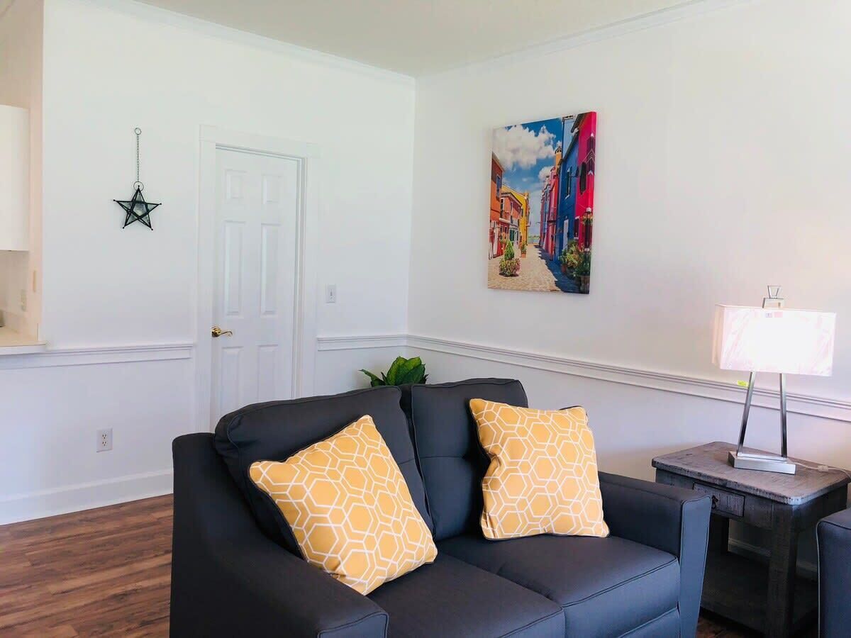A living room with a black leather sofa, two patterned cushions, a star-shaped wall decoration, a painting of a street scene, and a table lamp.