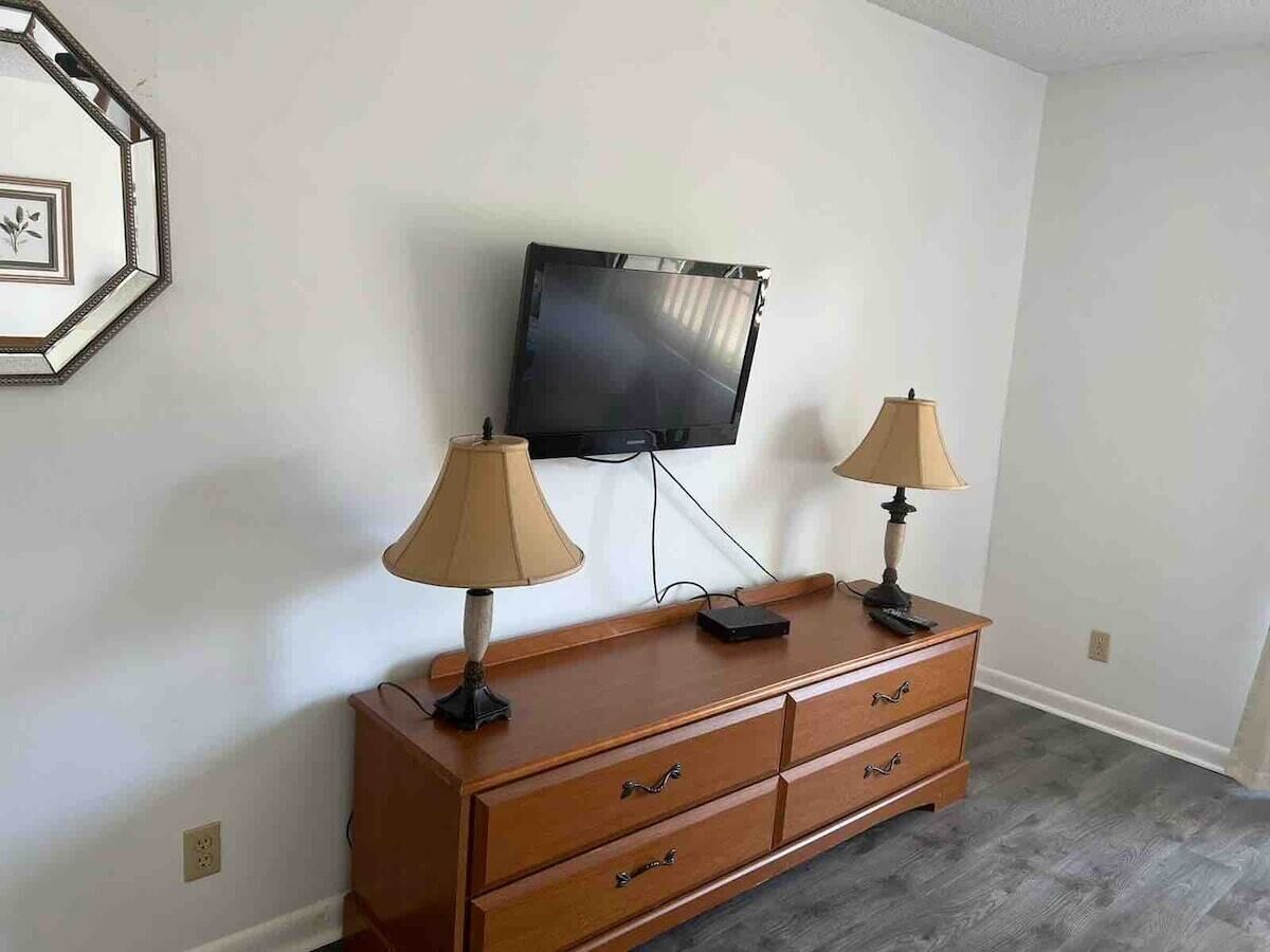 Une pièce comprenant une commode en bois, deux lampes, un téléviseur à écran plat fixé au mur et un tableau encadré accroché au mur.