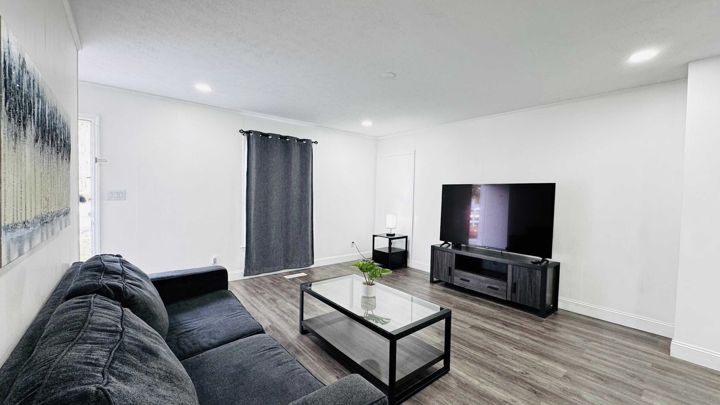 Uma sala de estar moderna com um sofá cinza escuro, uma mesa de centro de vidro, uma televisão de tela plana sobre um suporte de madeira e um quadro grande na parede.