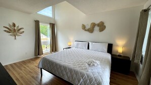 A bedroom with a large bed, two bedside tables with lamps, a window with curtains, and a wall decoration. - LR1 Bay Tree 1 bedroom condo with pool (Little River)