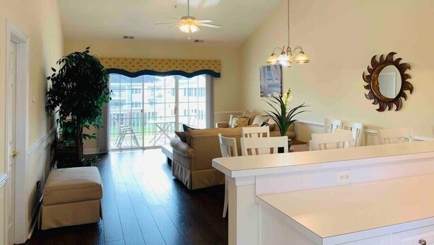 A spacious living room with a large window, a ceiling fan, and a dining area with a white table and chairs. - The perfect location for your vacation! (Myrtle Beach)
