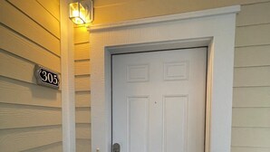 Puerta blanca con pomo y cerradura, enmarcada por un detalle blanco y una pequeña lámpara arriba. Se ve una placa con el número '305' en la pared a la izquierda.