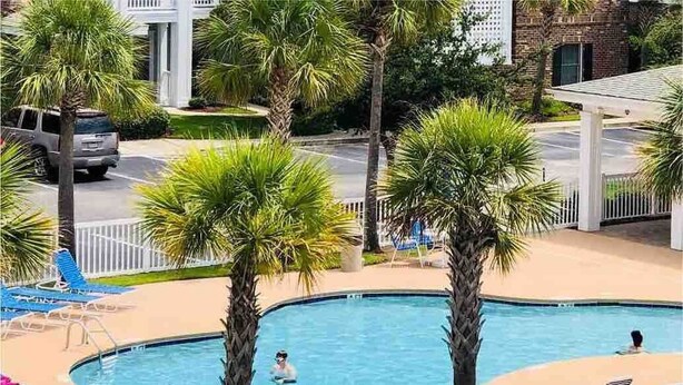 Zona de piscina con sillas de descanso, palmeras y un edificio al fondo.