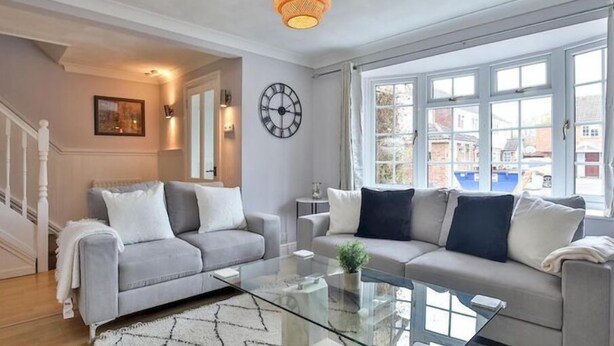 A living room with two sofas, a glass coffee table, a large window with a view, a wall clock, and a hanging light fixture. - haven Valley  near the theme park  (Old Windsor)