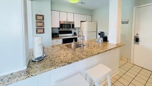 Una cocina con armarios blancos, encimera de granito, pileta de acero inoxidable y cafetera.