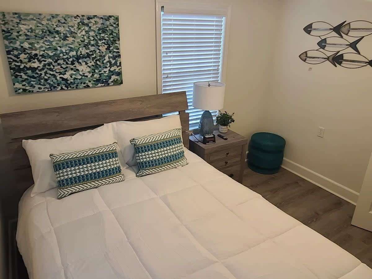 Een slaapkamer met een bed, twee bedkussens met een patroon, een nachtkastje met een lamp, een kleine plant en een kunstwerk aan de muur.