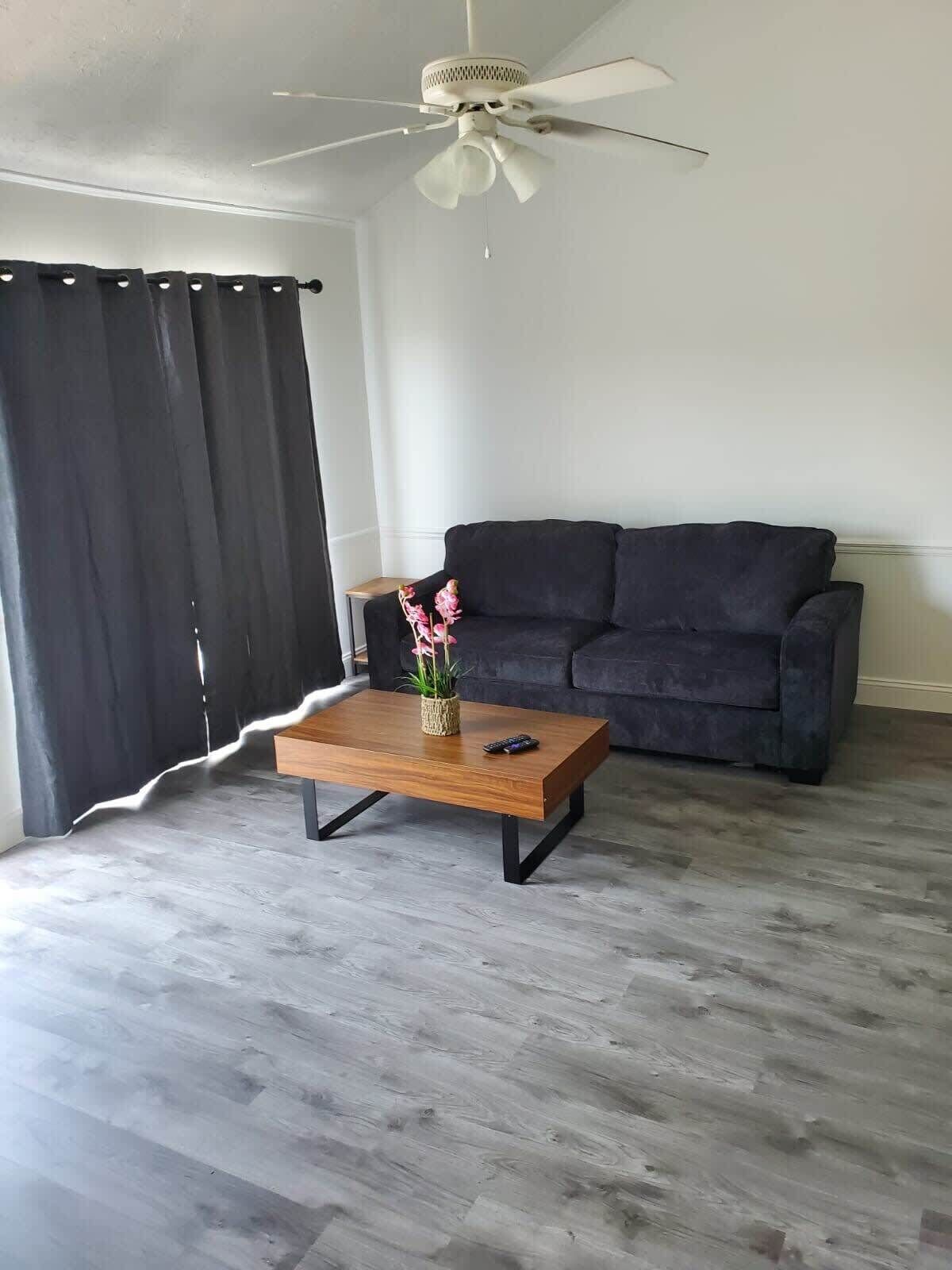 A living room with a dark sofa, a wooden coffee table with a vase and flowers, and a ceiling fan.