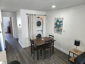Ein Essbereich mit Tisch und Stühlen, eine Waschmaschine im Hintergrund, ein Gemälde an der Wand und eine Lampe auf einem Beistelltisch.