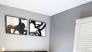Una cama bien hecha con colcha blanca, dos almohadas y un juego de toallas sobre la cama. Hay dos cuadros abstractos en la pared sobre la cama.