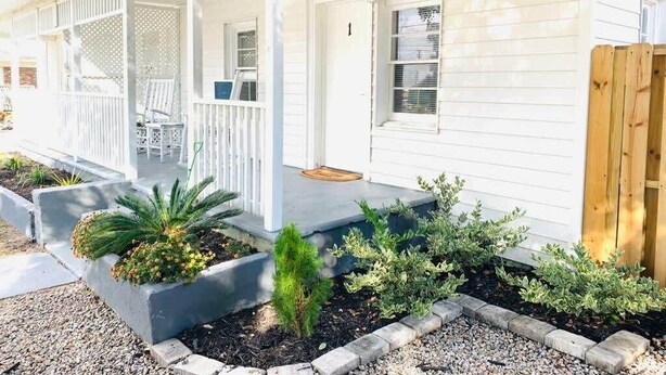 Una casa blanca con un porche cubierto, un pequeño jardín con diversas plantas y una cerca de madera.