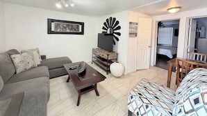 Una sala de estar con un sofá, una mesa de centro, un televisor y un adorno en la pared. Hay una puerta que conduce a un dormitorio.