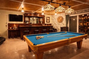 A pool table with balls, a bar area with stools, and a television mounted on the wall.