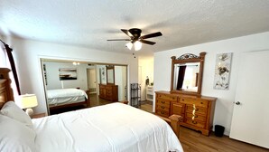Ein Schlafzimmer mit Bett, Schminktisch, Spiegel, Deckenventilator und einer Tür zu einem weiteren Raum.