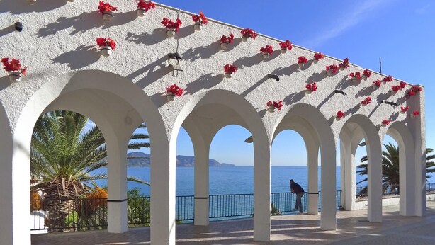Un paseo techado con arcos blancos y flores rojas, que conduce a una vista del mar.