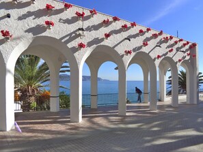 A covered walkway with white arches and red flowers, leading to a seaside view.