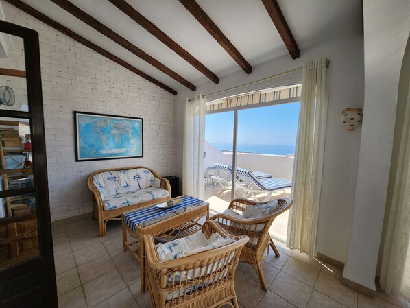 A room with a white brick wall, wooden beams, wicker furniture, a globe map, and a view of the sea.