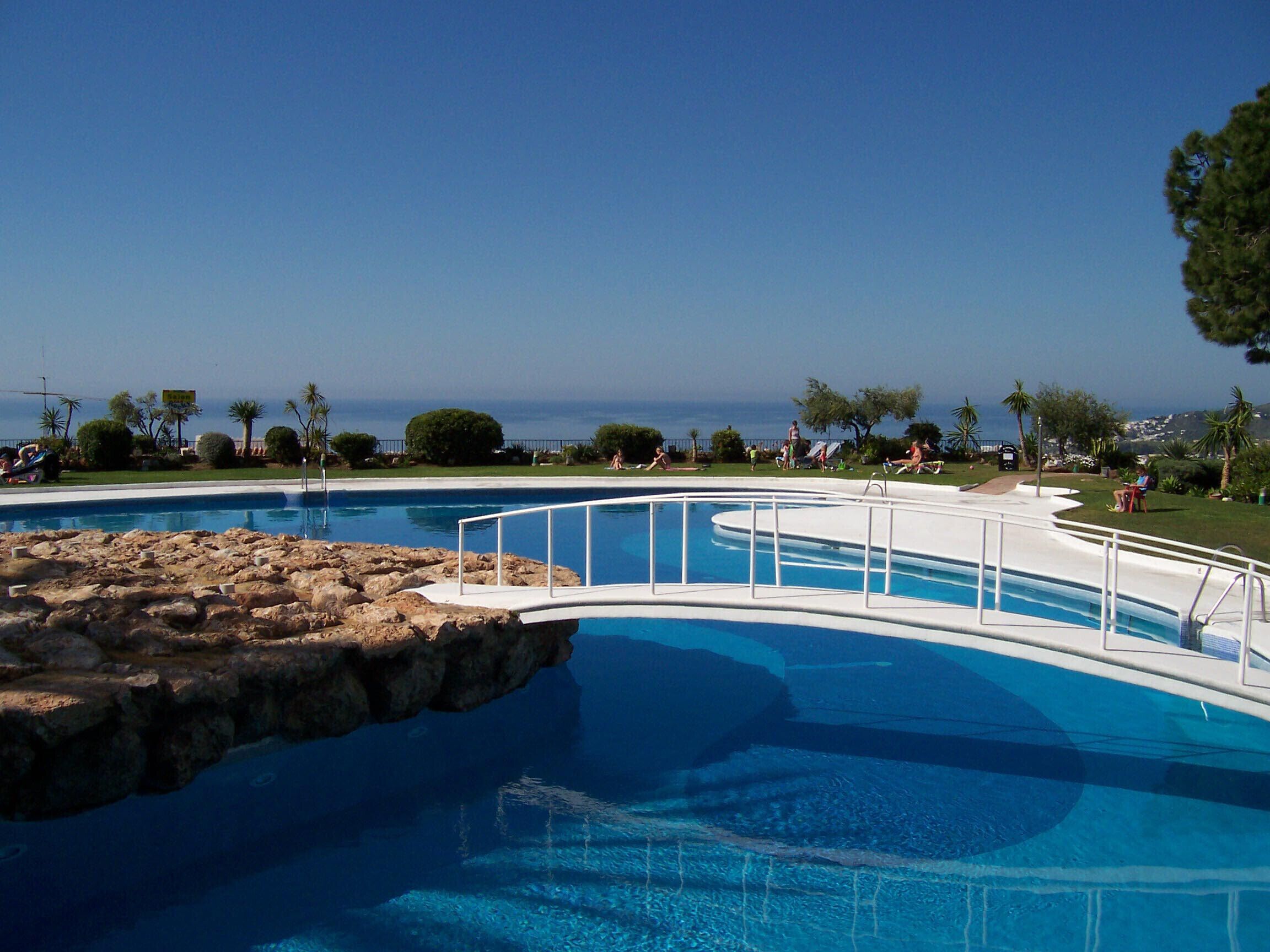 A pool with a white deck and a rocky edge, surrounded by greenery and a clear view of the ocean.