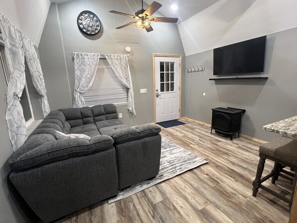 A modern living room with a sectional sofa, a ceiling fan, a fireplace, and a flat-screen TV mounted on the wall. - Quaint cozy cabin near Hatcher Pass. Clear Night Cabin B (Wasilla)
