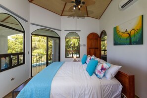 A bedroom with a large bed, a ceiling fan, and a view of the outdoors through arched windows.