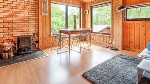 A wooden cabin interior with a brick wall, a fireplace, a dining table with chairs, and large windows offering a view of greenery. - 2 bedroom accommodation in Kilmorack, near Beauly (Kilmorack, near Beauly)