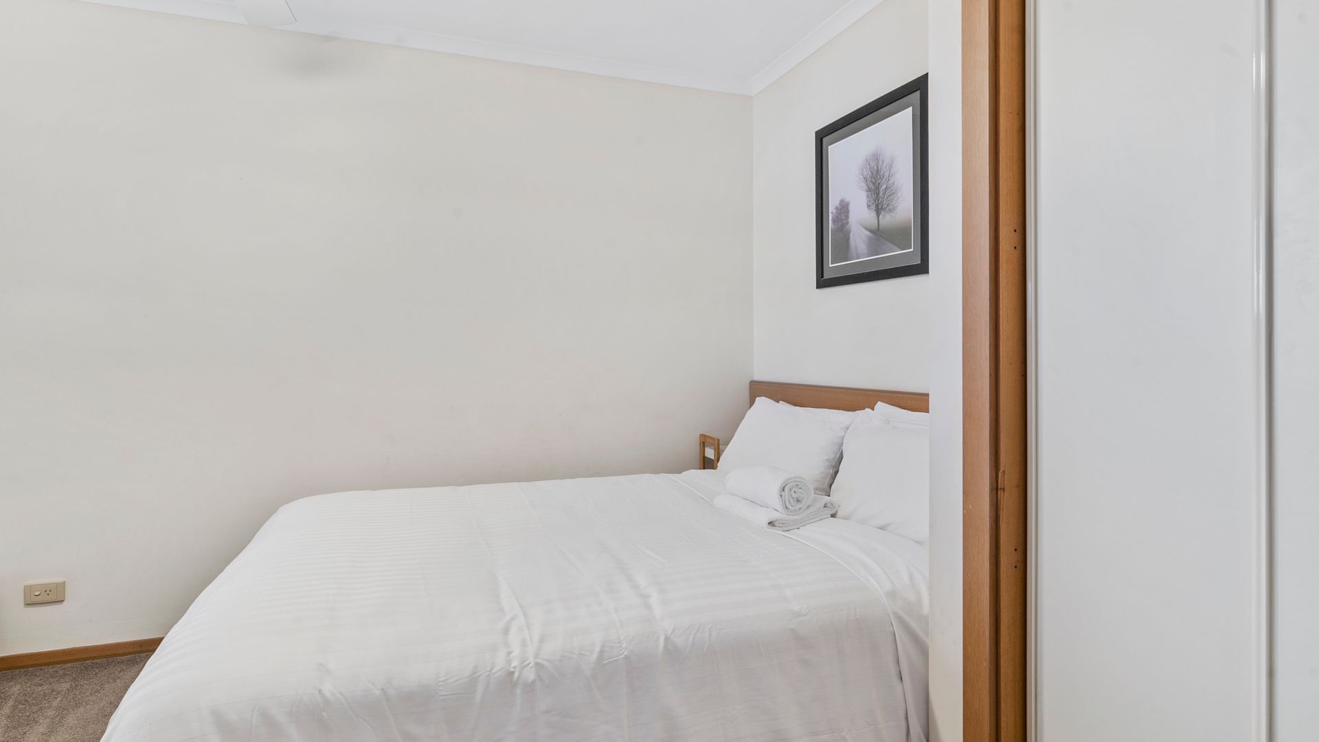 Cama bem arrumada em quarto com paredes brancas, um quadro na parede e moldura de porta de madeira.