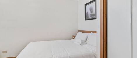 Cama bem arrumada em quarto com paredes brancas, um quadro na parede e moldura de porta de madeira.