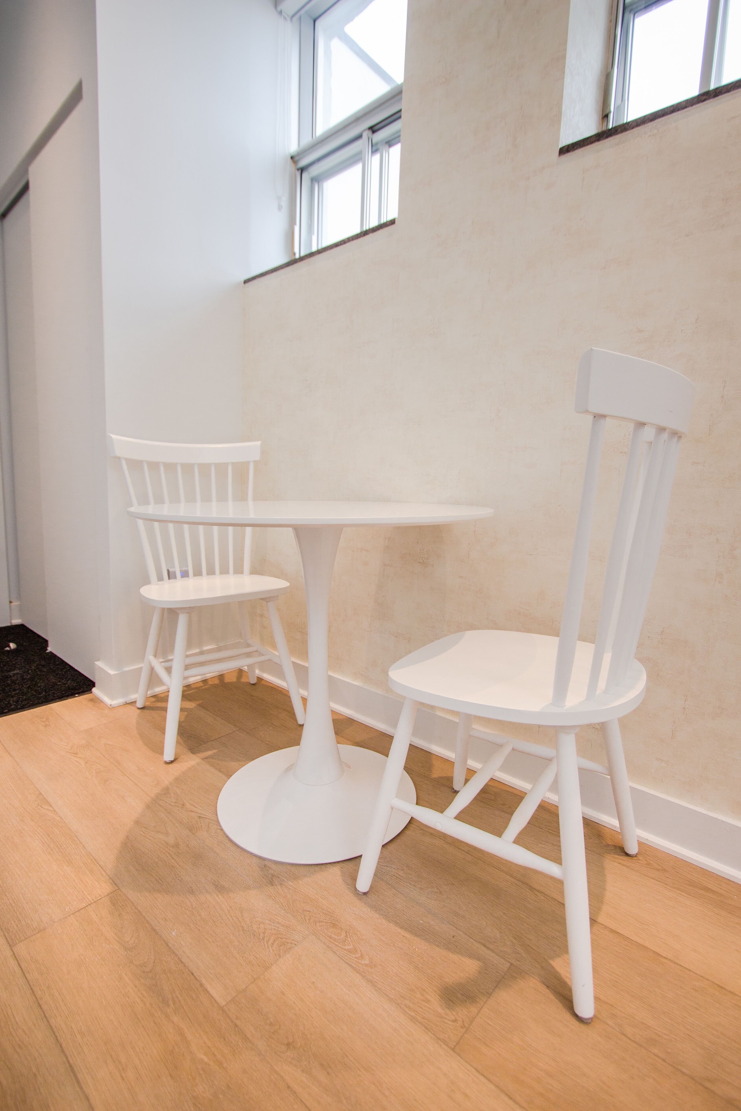 A corner of a room with a white table and two white chairs, a black doormat, and a wooden floor.